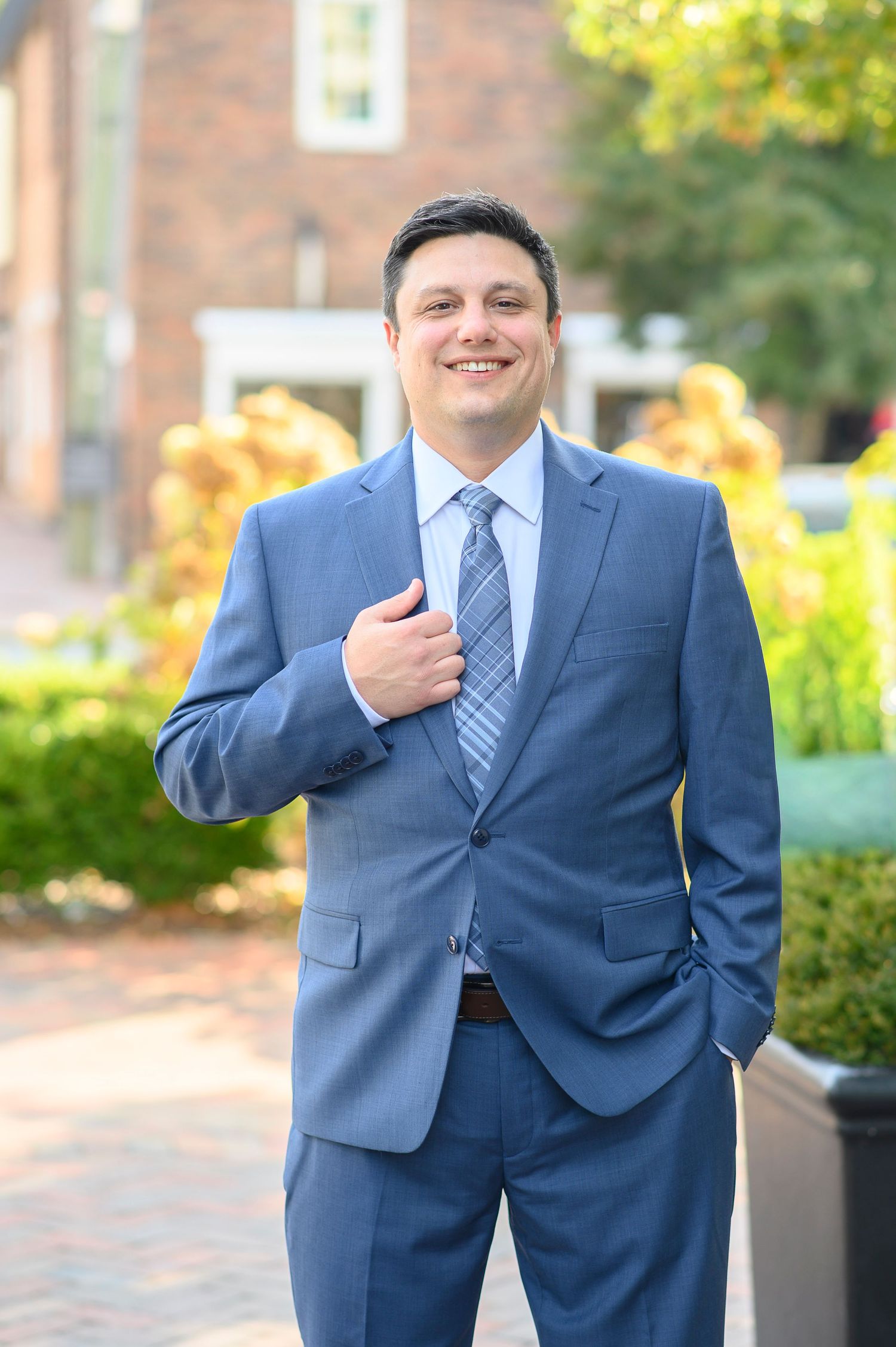 groom in a navy blue suit and tie stands outdoors near landscaping and brick buildings. wedding photos in alpharetta, GA