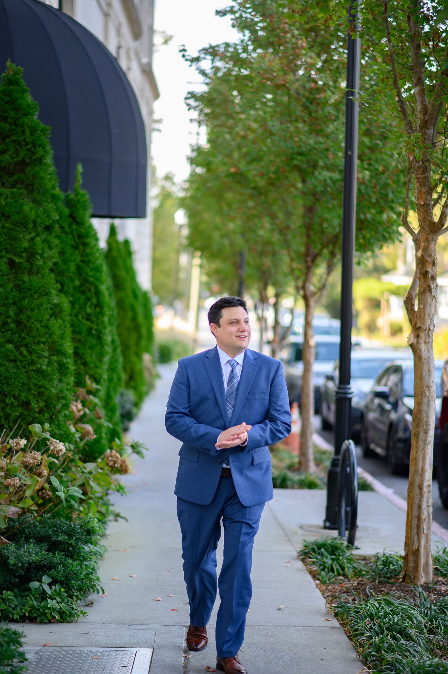 A groom in a navy blue suit walks and poses along a tree-lined urban sidewalk in a business district. wedding photos in alpharetta, GA