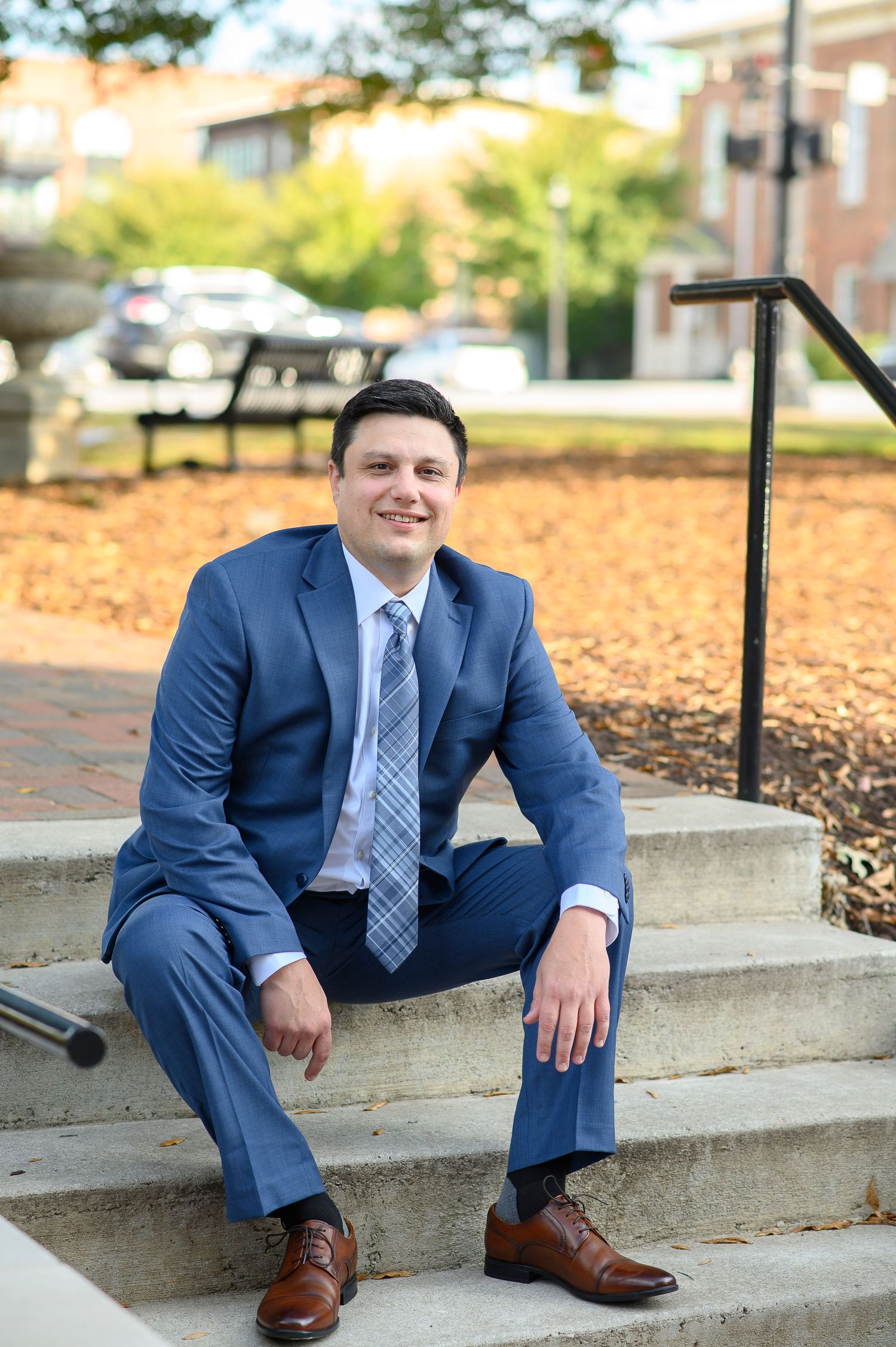 A person in a navy blue suit and striped tie sits casually on concrete steps surrounded by autumn leaves. wedding photos in alpharetta, GA