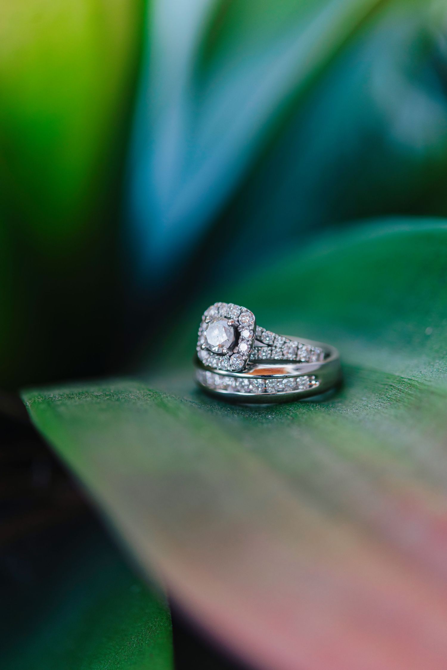 Sparkling diamond engagement ring and wedding band set photographed with artistic bokeh on green leaf background.