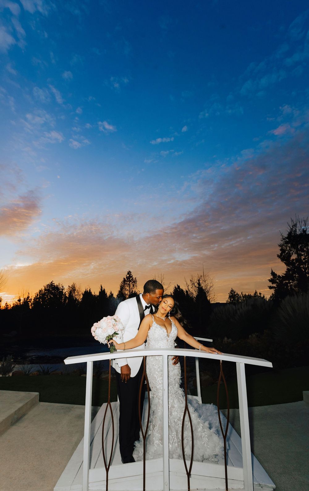 A couple in wedding attire shares romantic moments at sunset against a blue sky and silhouetted trees.