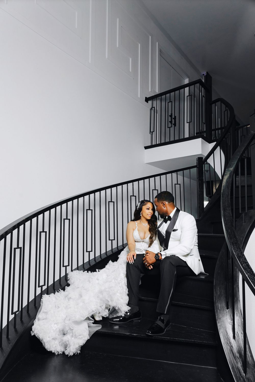 A bride in a flowing white dress and groom in white jacket sit intimately on elegant curved black staircase.
