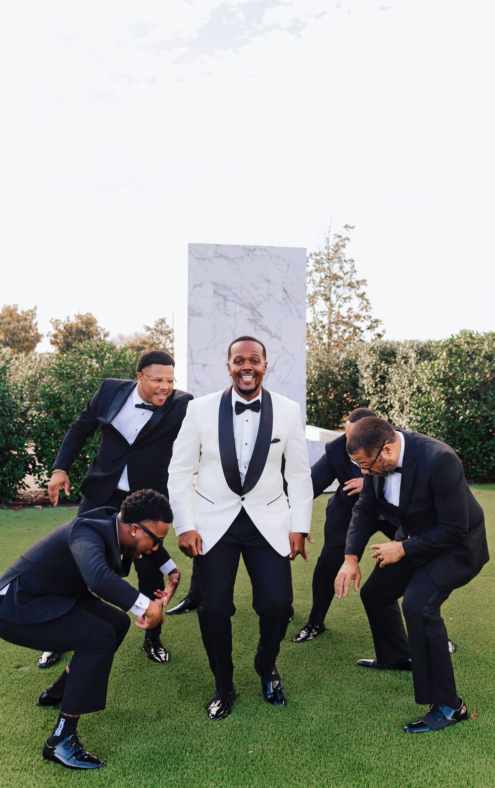 Groomsmen in black and white formal attire playfully pose on grass in a sequence of fun wedding party photos.