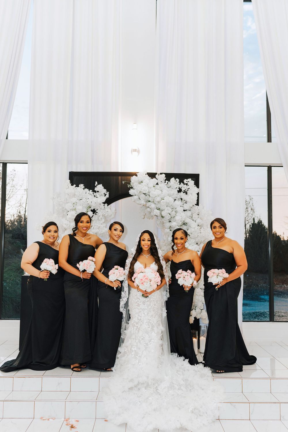 Bridesmaids in black dresses pose with bride in white against draped backdrop.