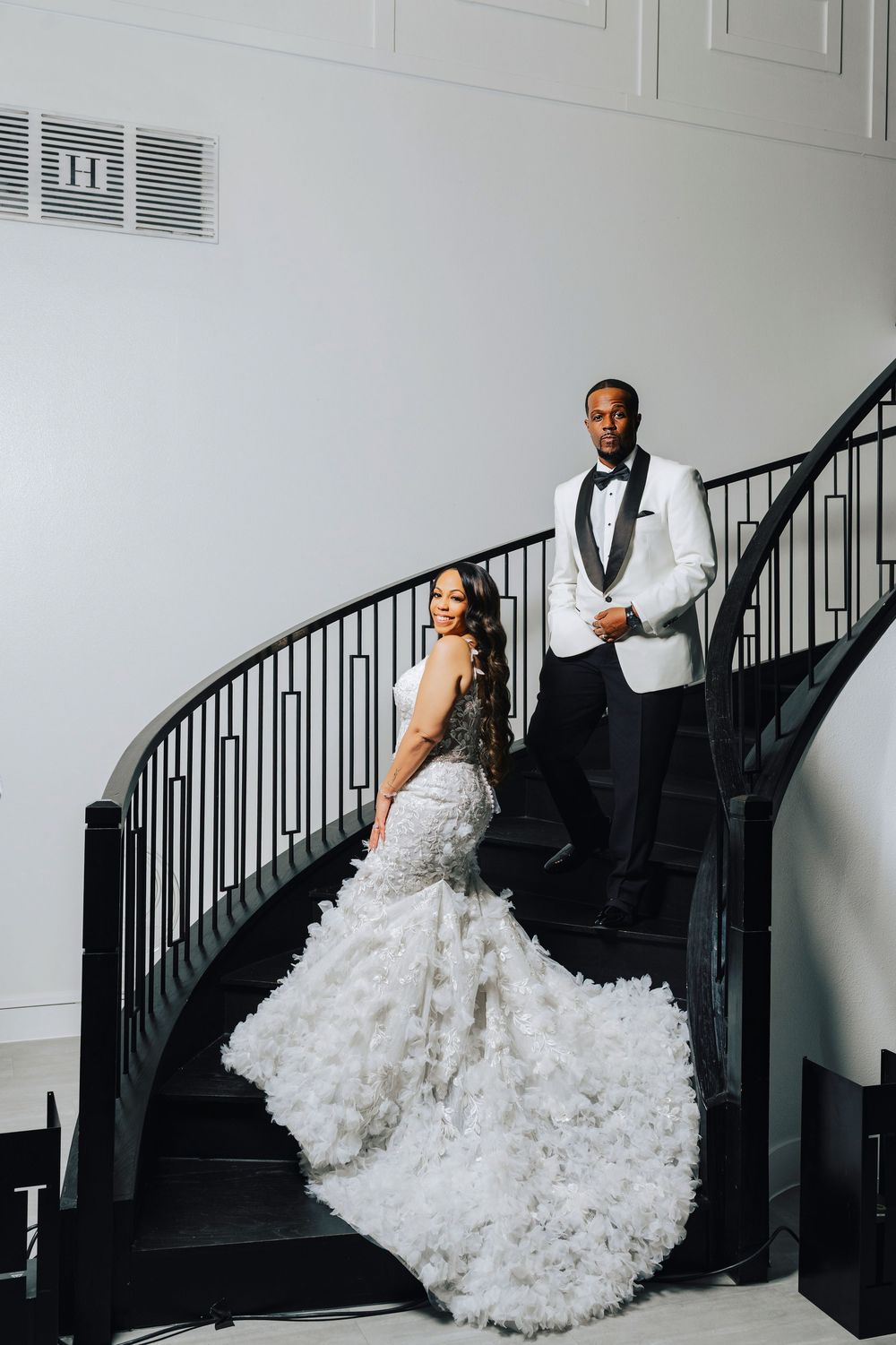 A bride in a ruffled white gown and groom in formal attire pose dramatically along an elegant black staircase.
