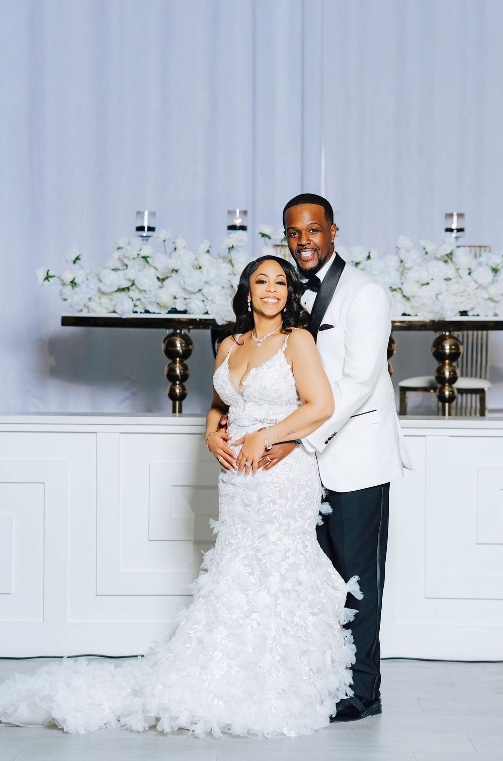 A wedding couple shares intimate moments in an elegant white room with floral decorations.