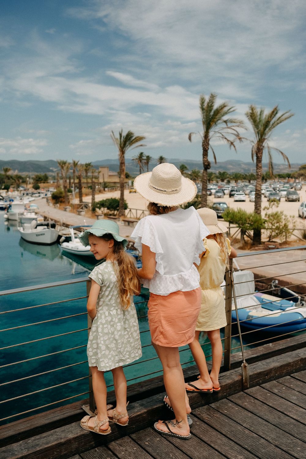 People in summer hats enjoying a view of boats and palm trees from a wooden dock at a marina.