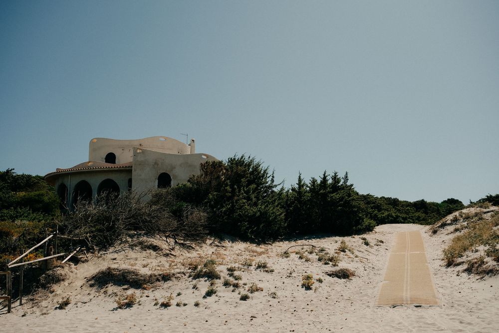 Sandy beach pathway leading to a Mediterranean style building on a coastal hillside.