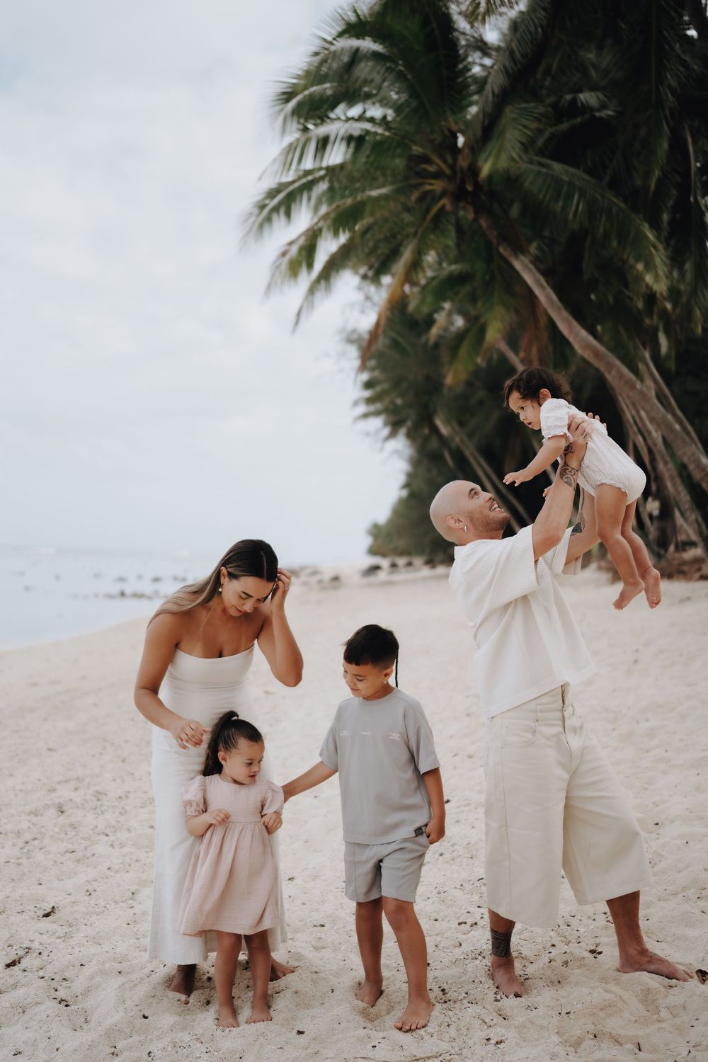 A family in coordinated white and beige outfits walks together on a tropical beach with palm trees in the background.