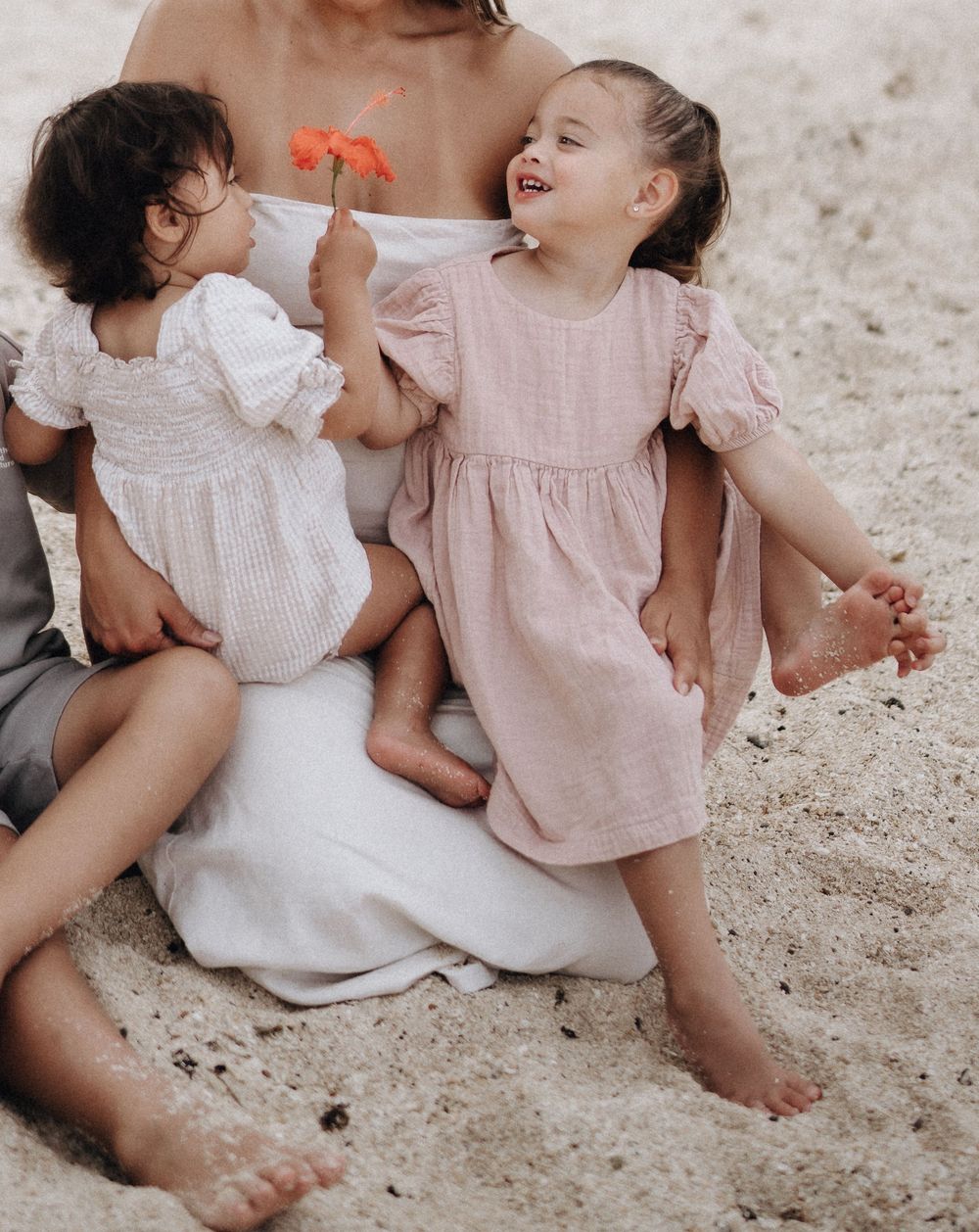 Children sitting together on sandy ground wearing white and pink dresses in a candid summer scene.