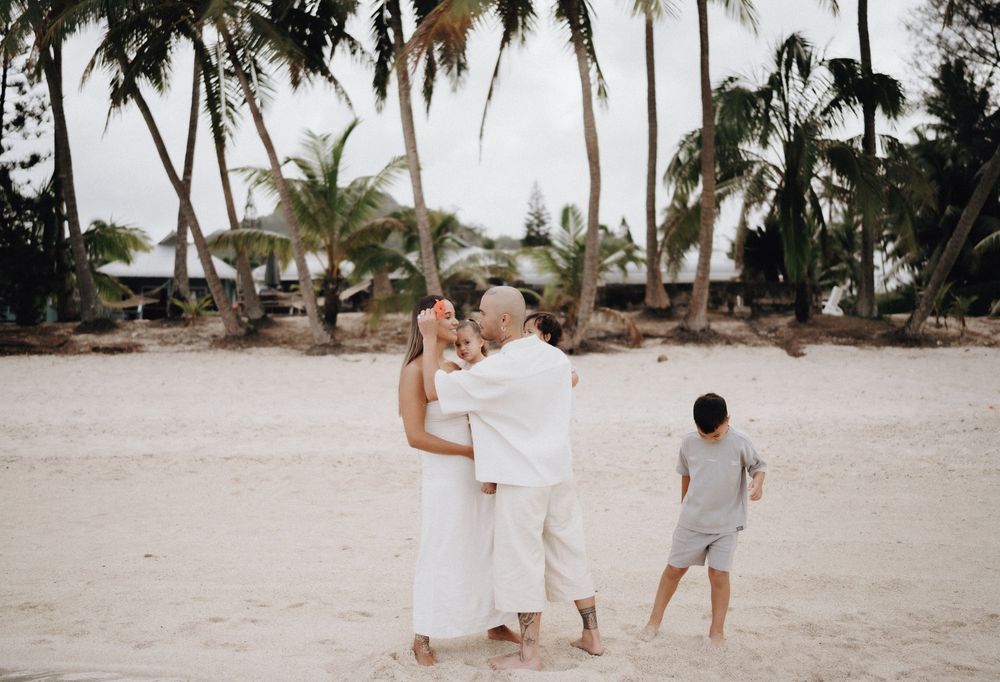 A couple in white outfits walks hand in hand along a tropical beach with palm trees and ocean views.