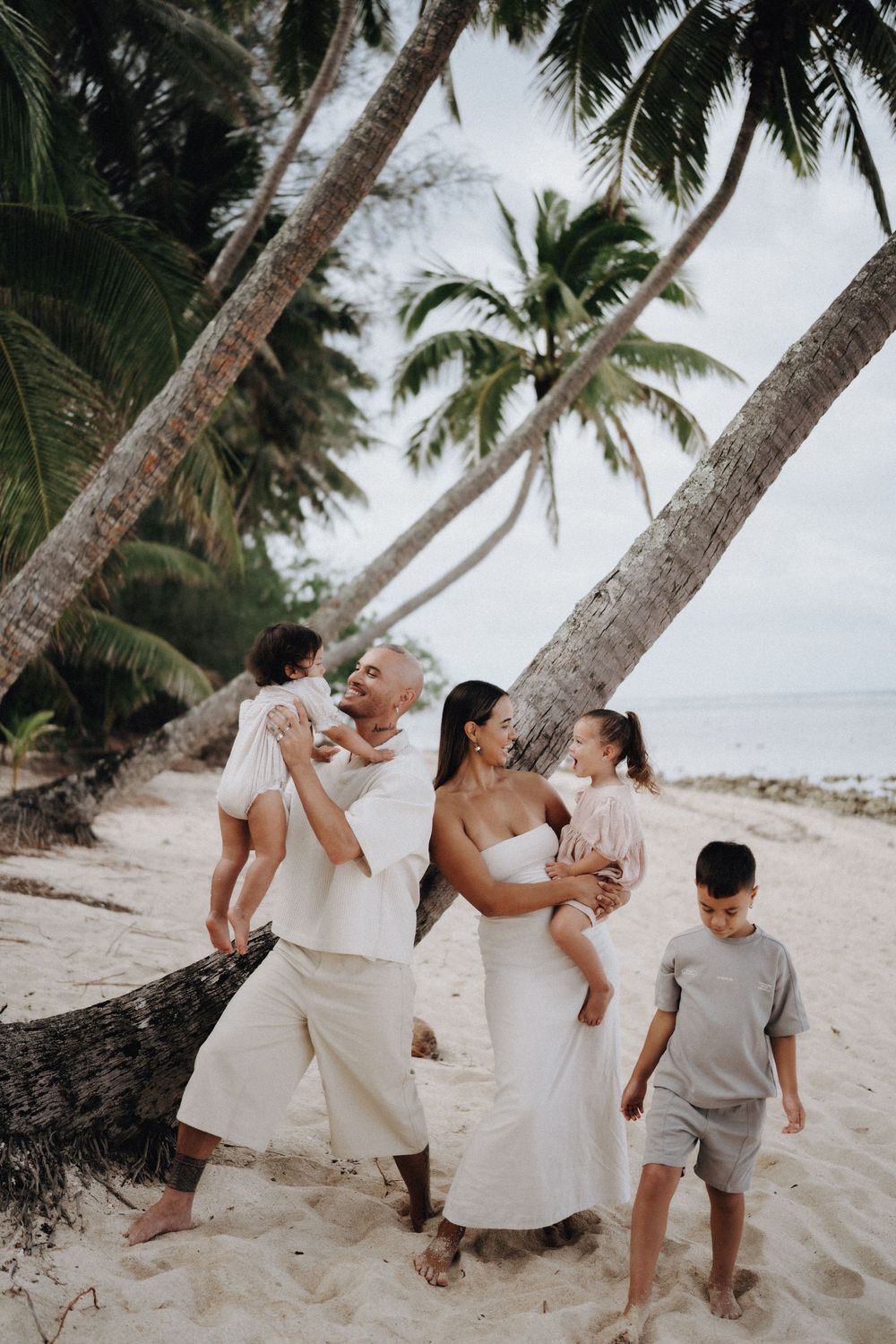 A family shares joyful moments together on a tropical beach with palm trees and white sand in the background.