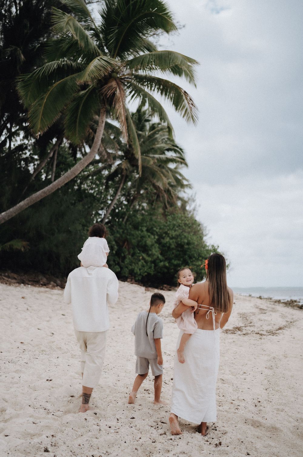 Family walking together along a tropical beach with palm trees and cloudy skies in the background.