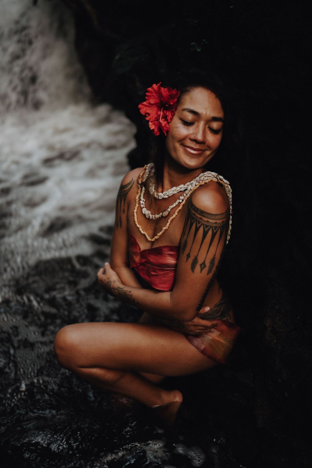 A woman in a red dress with traditional jewelry and flowers sits on dark rocks amid atmospheric mist.