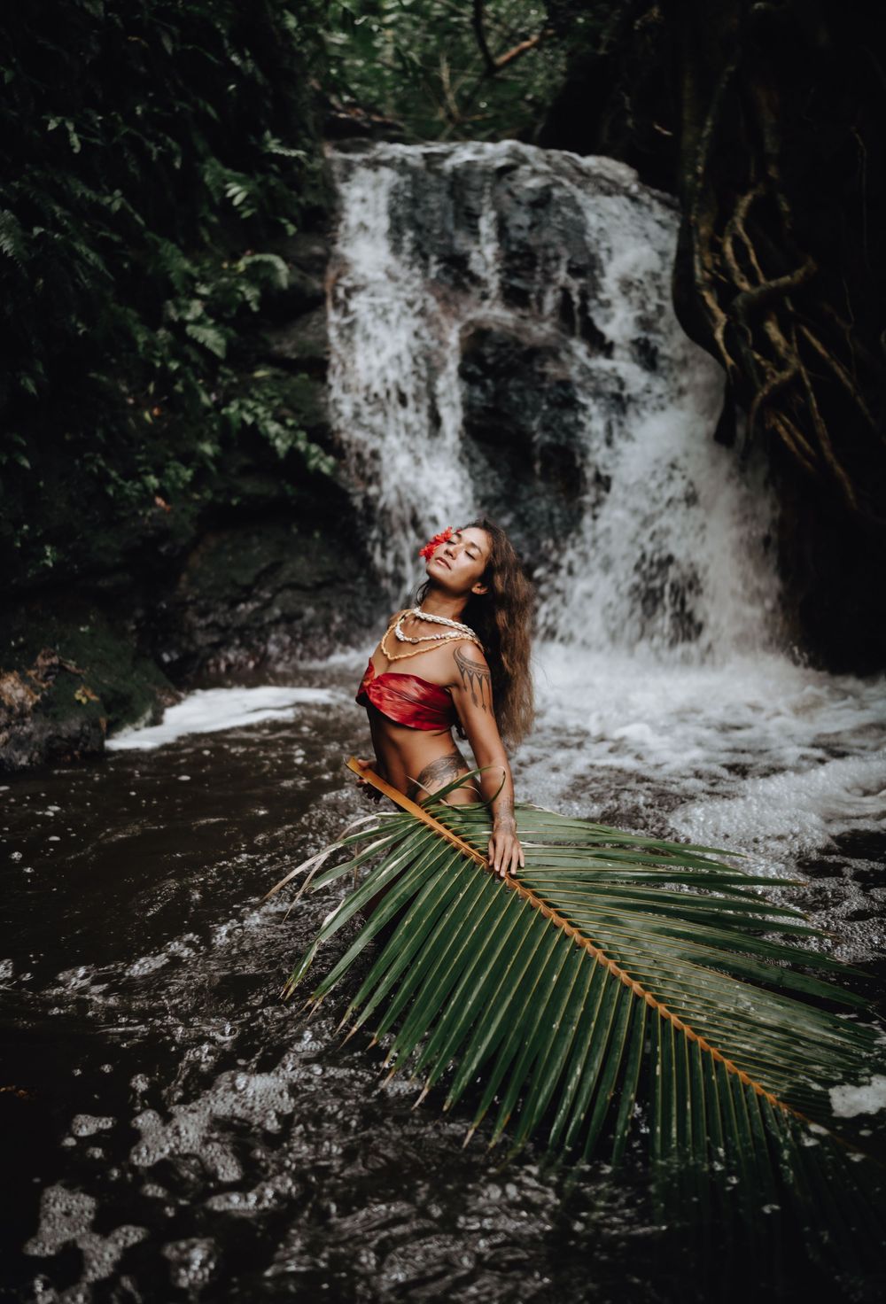 A tropical photoshoot by a waterfall features a palm frond and red dress against dark rocks and cascading water.