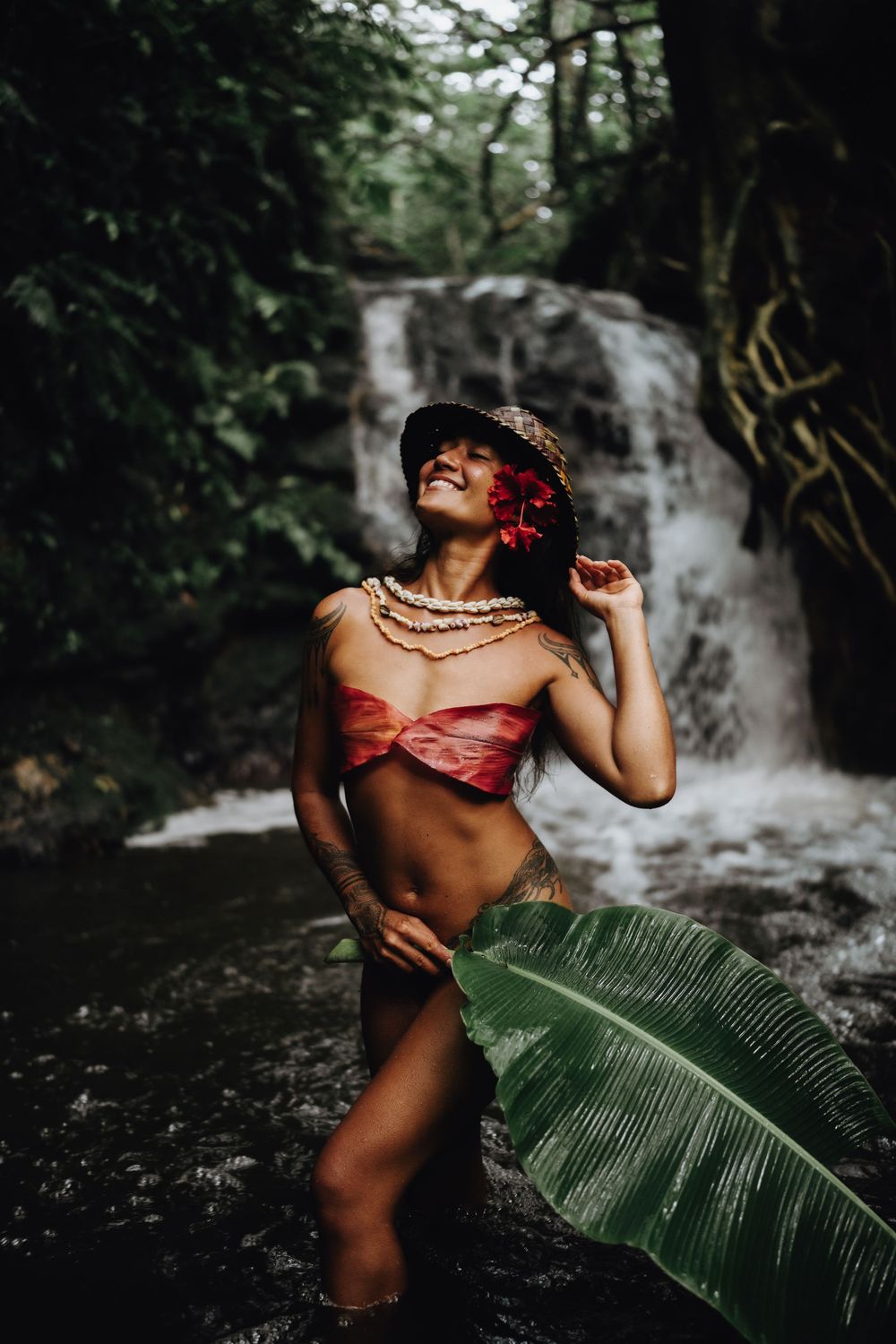 Artistic tropical photoshoot at a waterfall featuring traditional Hawaiian-inspired styling with red top and palm frond.