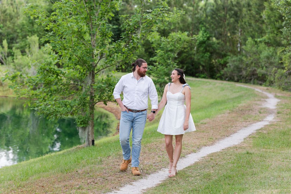 A couple strolls together along a peaceful dirt path next to a tranquil pond surrounded by lush greenery.