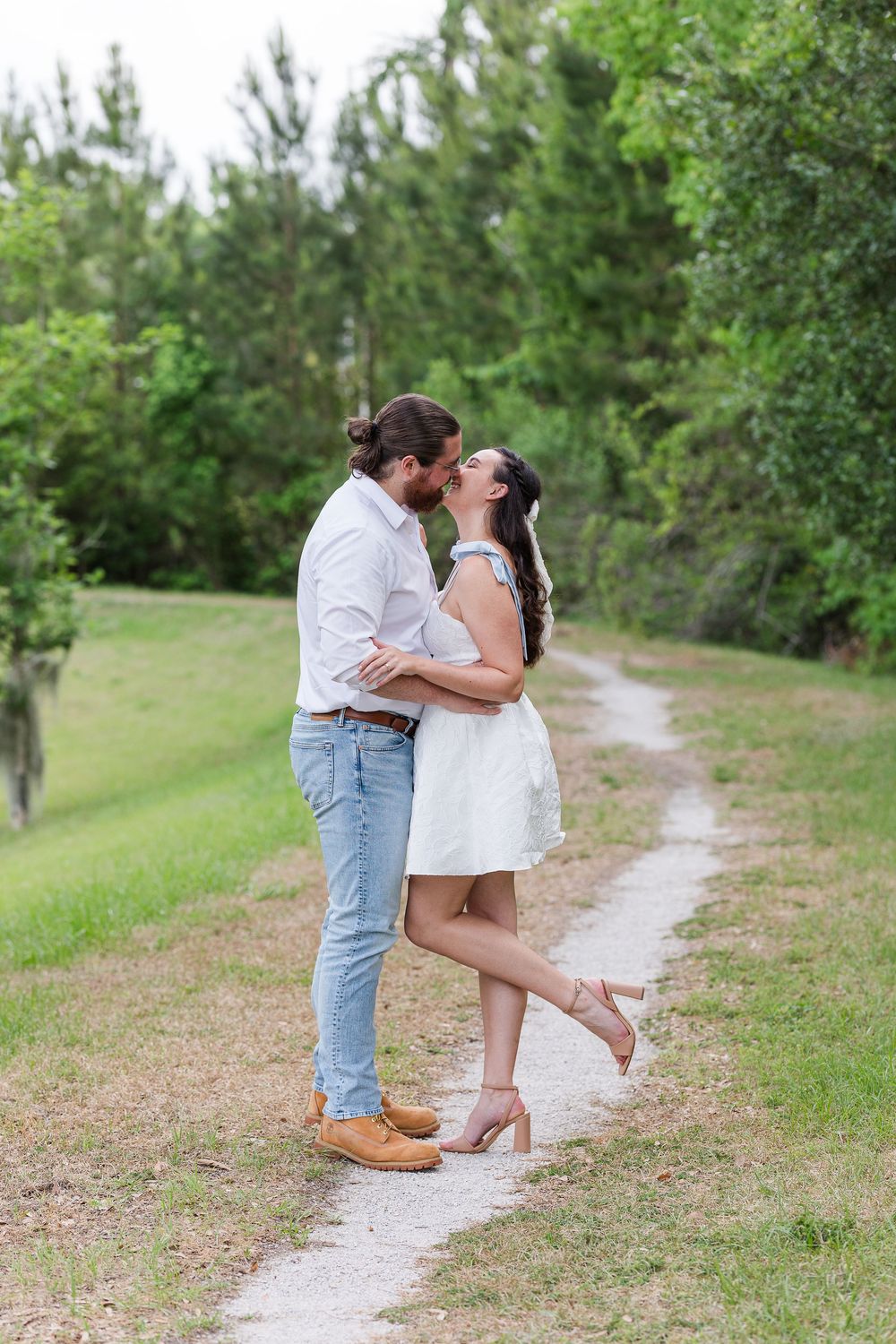 A romantic couple shares an intimate moment on a winding path amidst green trees and natural surroundings.