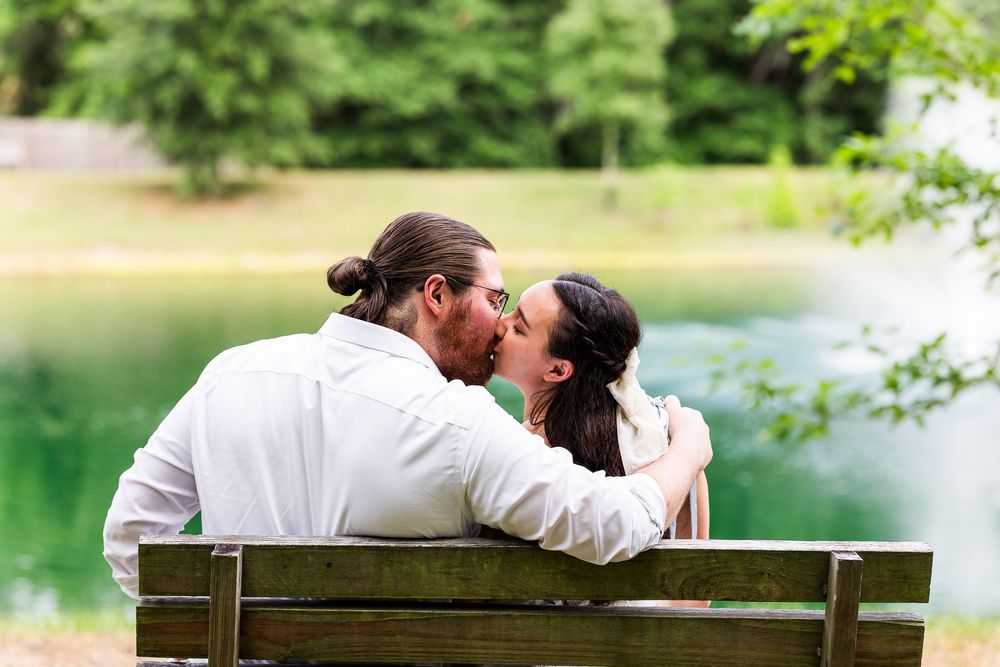 Romantic couple sharing tender moments on a park bench by a serene lake surrounded by lush greenery.