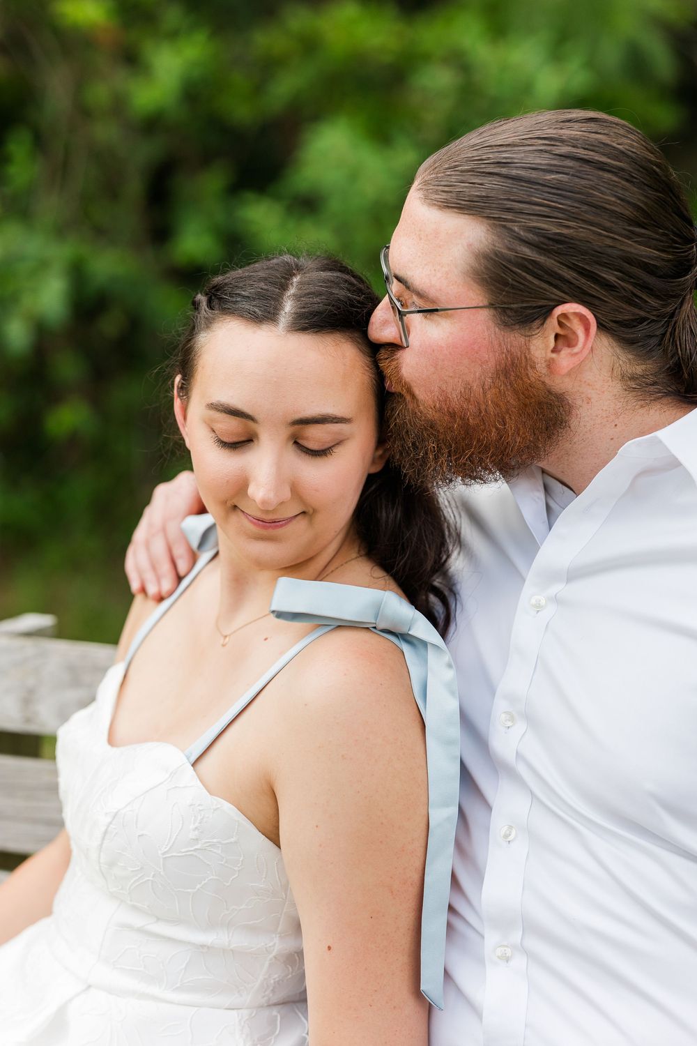 Intimate moment captured between couple in white and light blue attire against natural background.