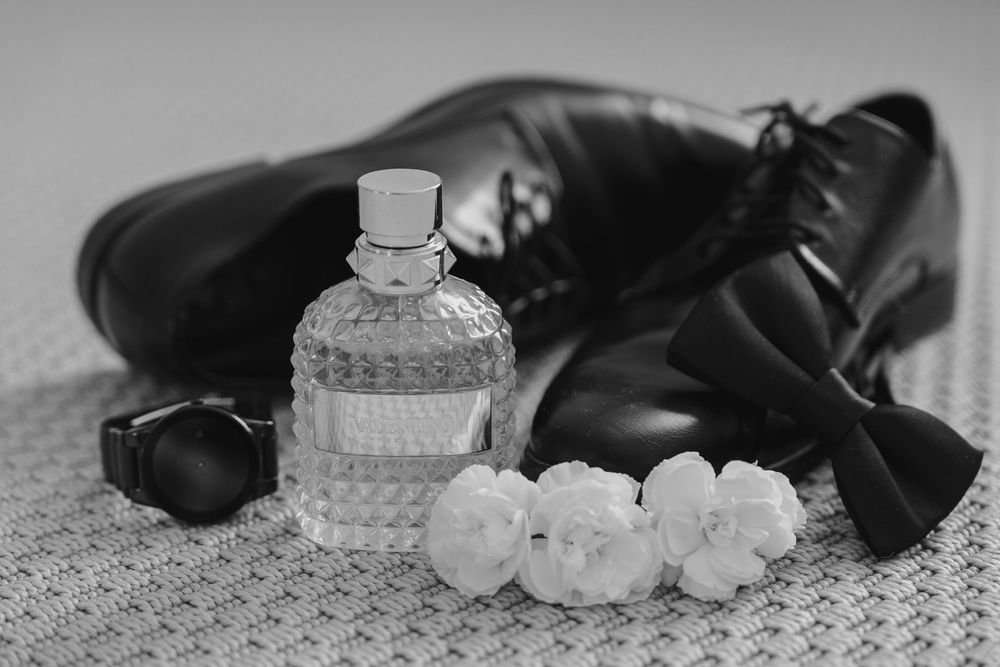 Black and white photo of formal dress shoes with perfume bottle and white floral decorations on lace tablecloth.