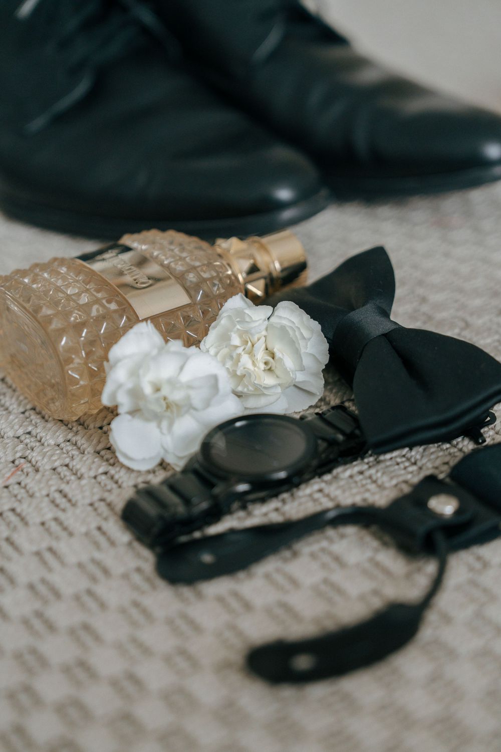 Black dress shoes and white floral decorations arranged on a textured beige surface.