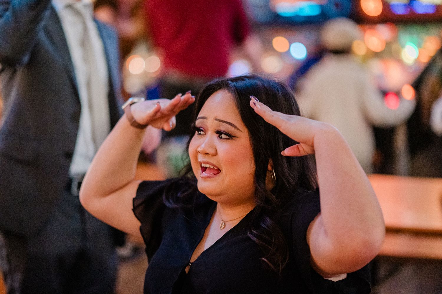 A person in a black dress dances joyfully at a party with blurred lights in the background.