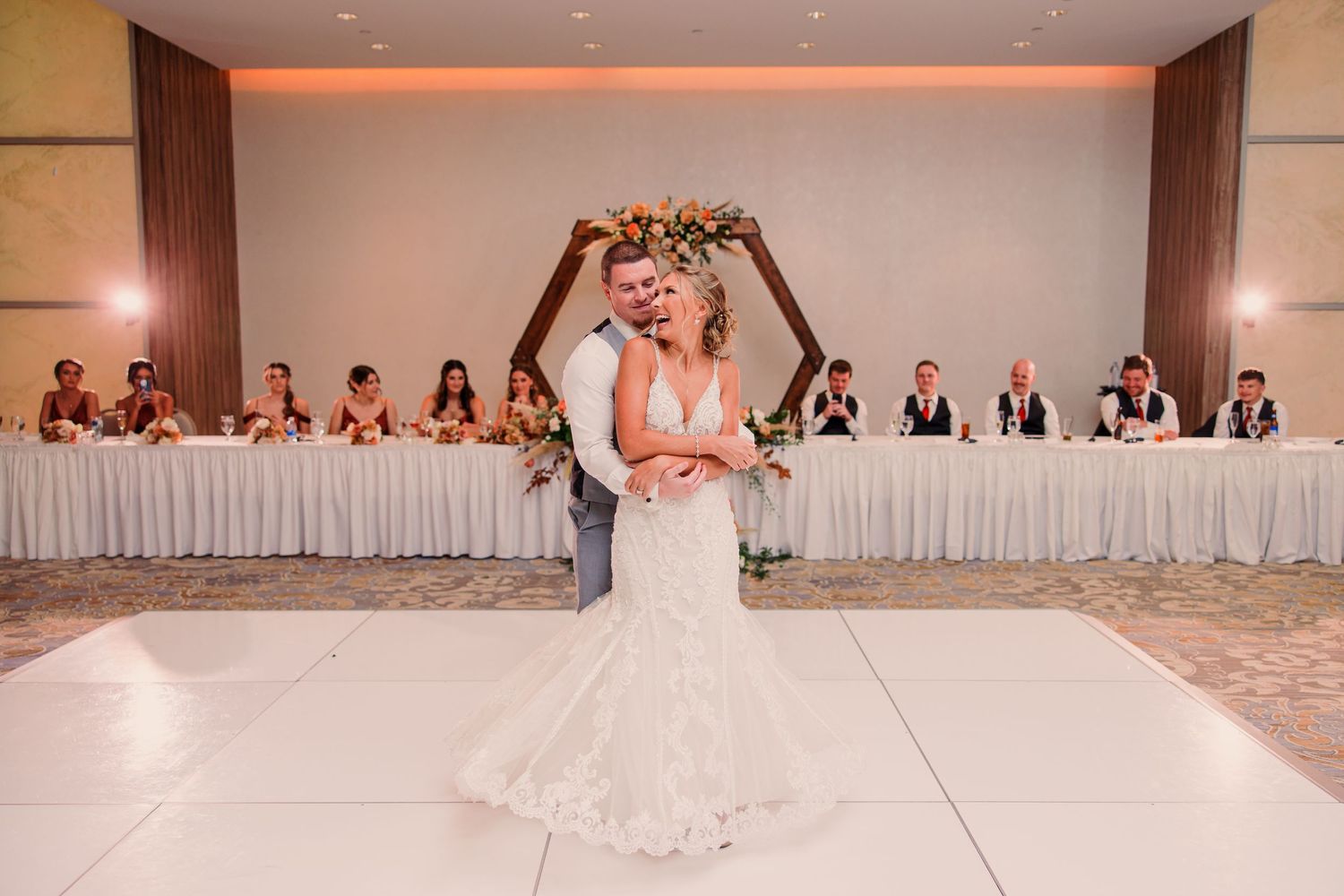 A bride in a lace wedding gown holds her bouquet on a white dance floor with wedding party seated at tables behind her.