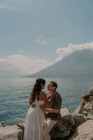 A couple shares an intimate moment on rocky shores with a scenic mountain and lake view in the background.