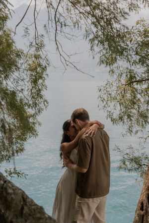 A couple embraces near a lake with willow tree branches hanging overhead in a romantic outdoor setting.
