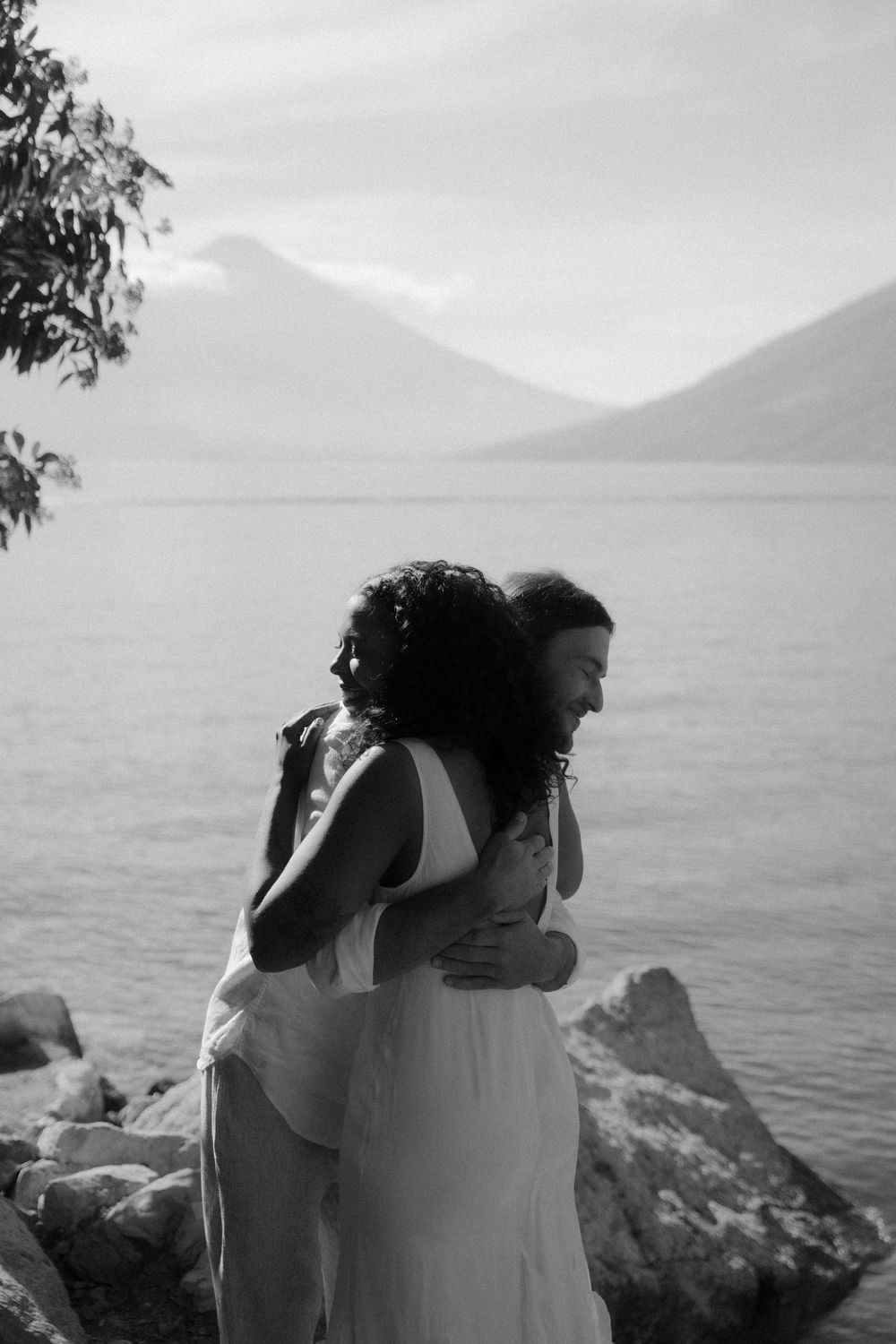 A romantic black and white photo series showing a couple embracing by the ocean with mountains in the background.