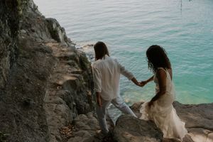 Two people holding hands walk along a rocky coastal cliff overlooking bright turquoise ocean waters.