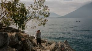 A panoramic view of a rocky coastline with turquoise waters and mountains in the distance under cloudy skies.