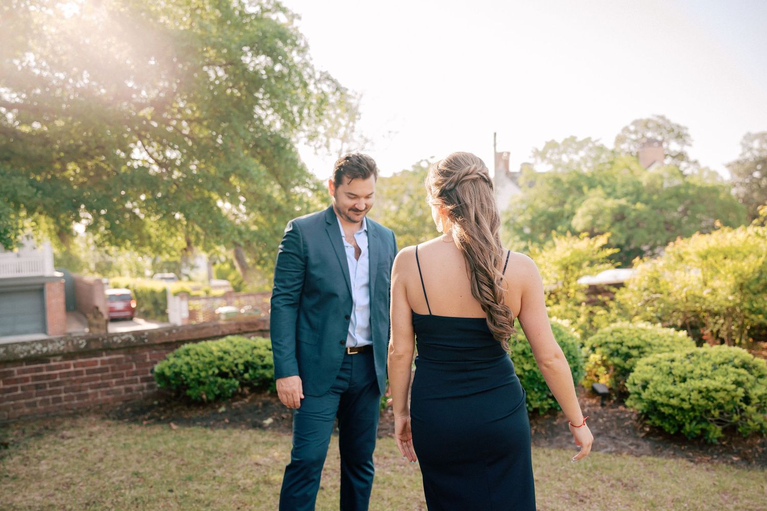 Couple in elegant attire walking outdoors, man in teal suit smiling at woman in black dress, lush greenery background.
