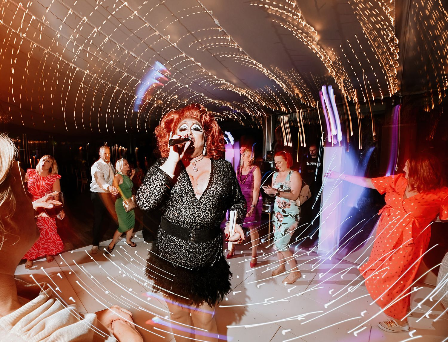 Drag queen performer singing on stage with microphone in a glittering tunnel-lit nightclub venue.