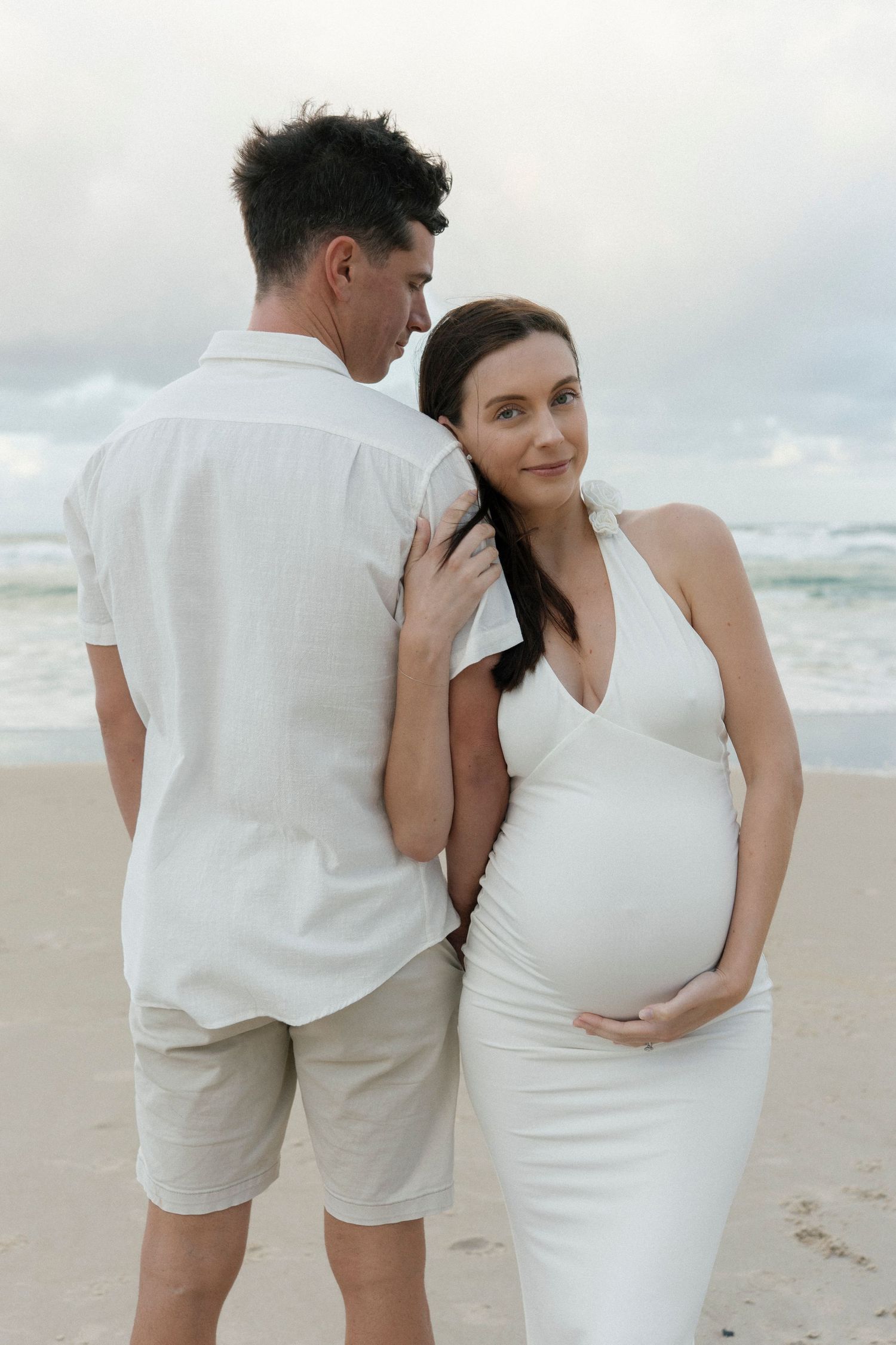 Pregnant woman in white dress with partner on beach, maternity photoshoot by the ocean.