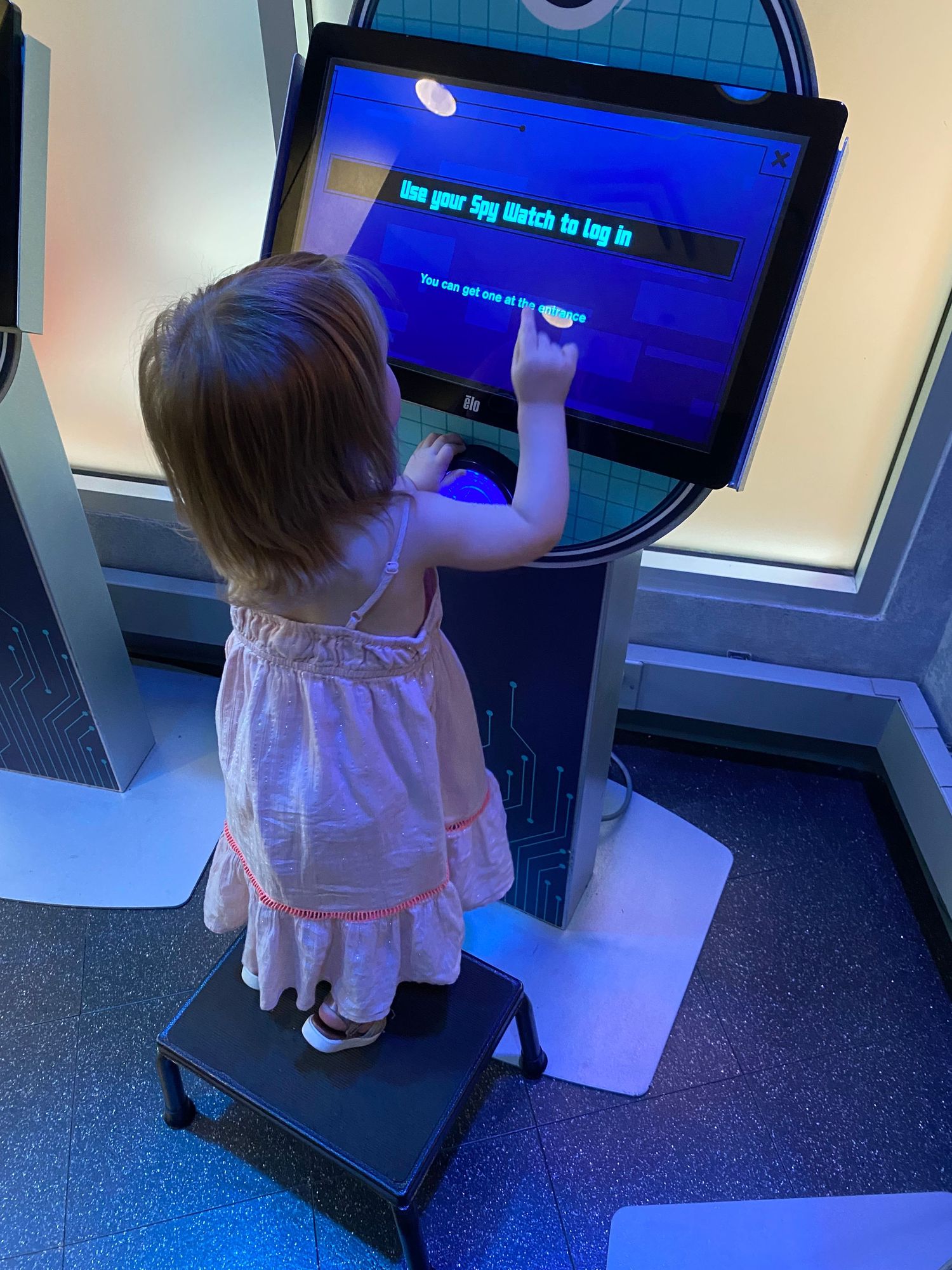 Toddler girl in pink dress standing on step stool using interactive touchscreen kiosk at children's museum.