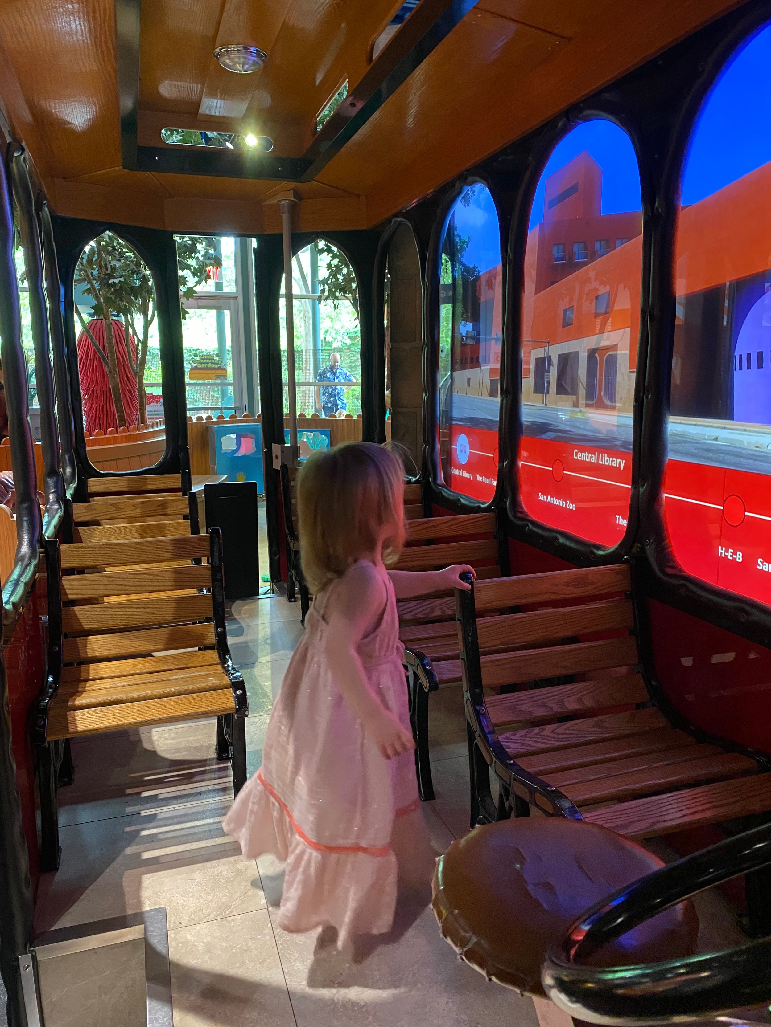 A toddler girl in a pink dress stands inside a vintage trolley car with wooden bench seats and scenic windows.