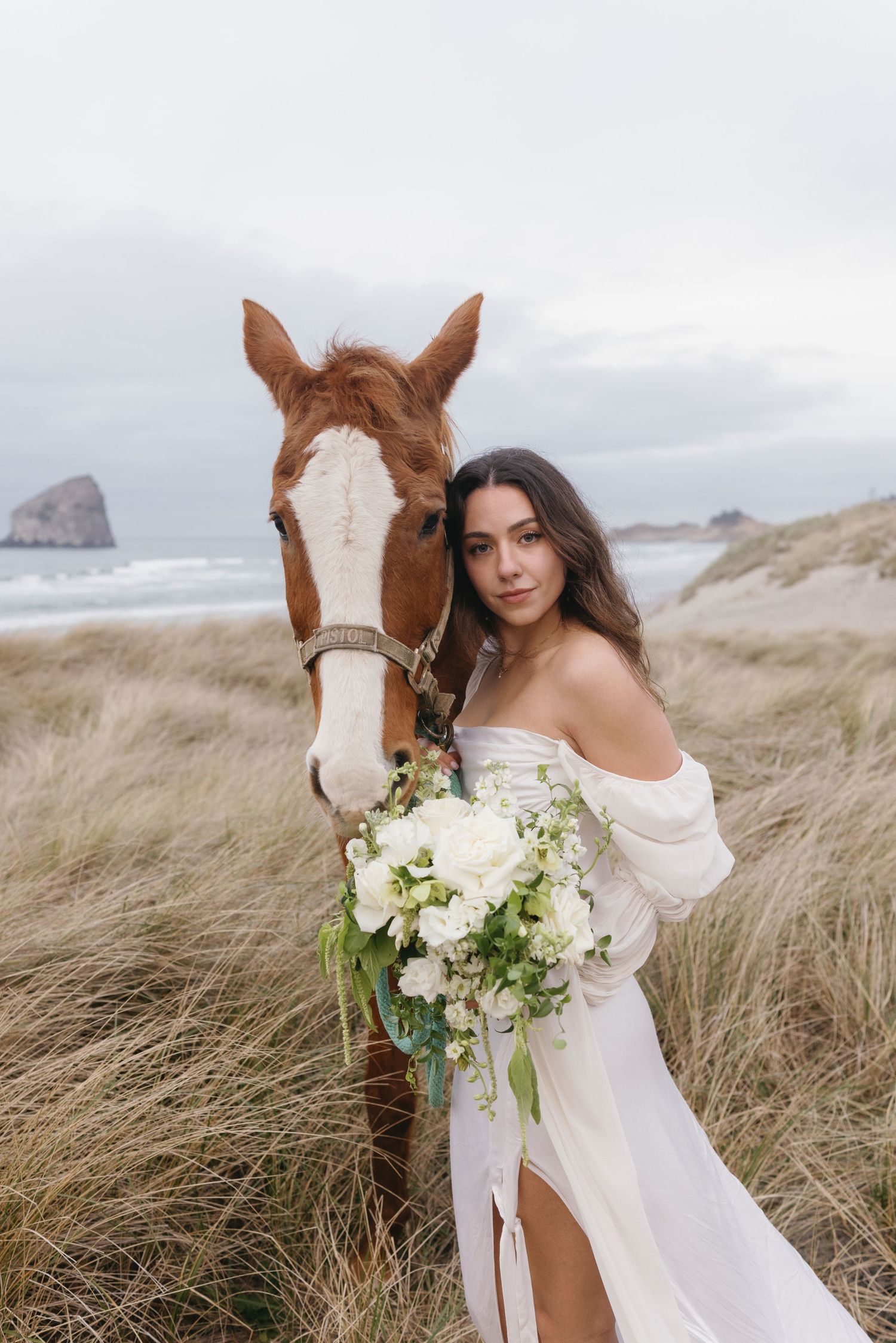 Bride in white off-shoulder gown holds white bouquet beside chestnut horse on a sandy beach.