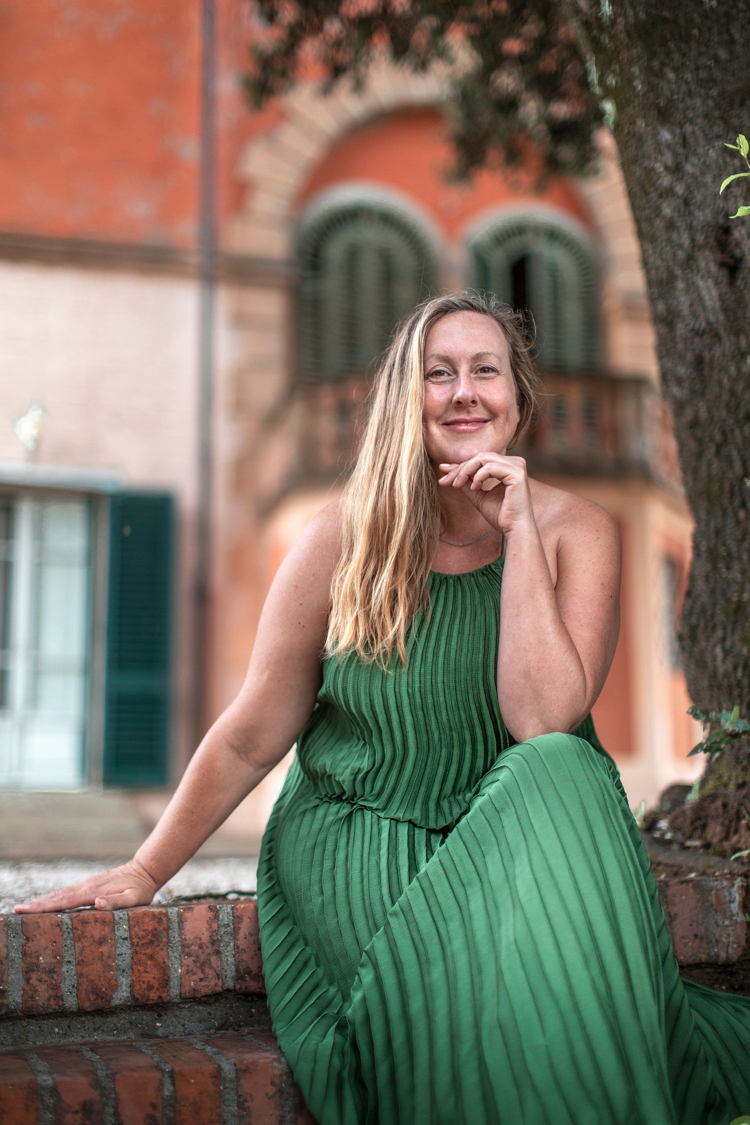 A smiling blonde woman in a green pleated dress sits relaxed in front of a rustic Italian villa.
