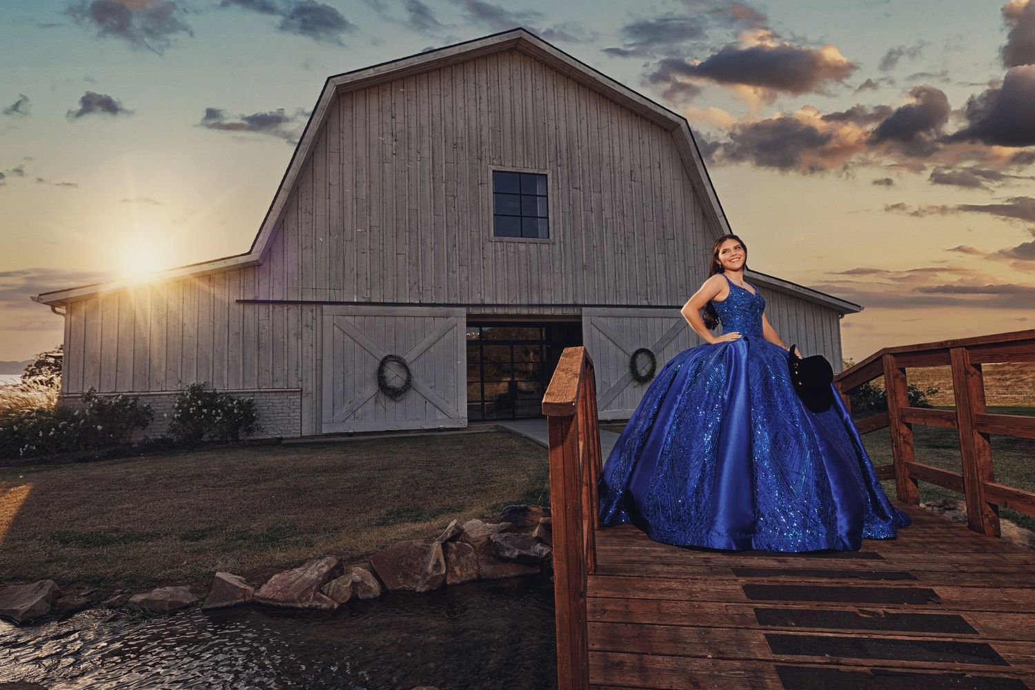 A girl in a sparkling royal blue Quinceañera gown stands on a wooden bridge in front of a white rustic barn at sunset in Carrollton, TX.