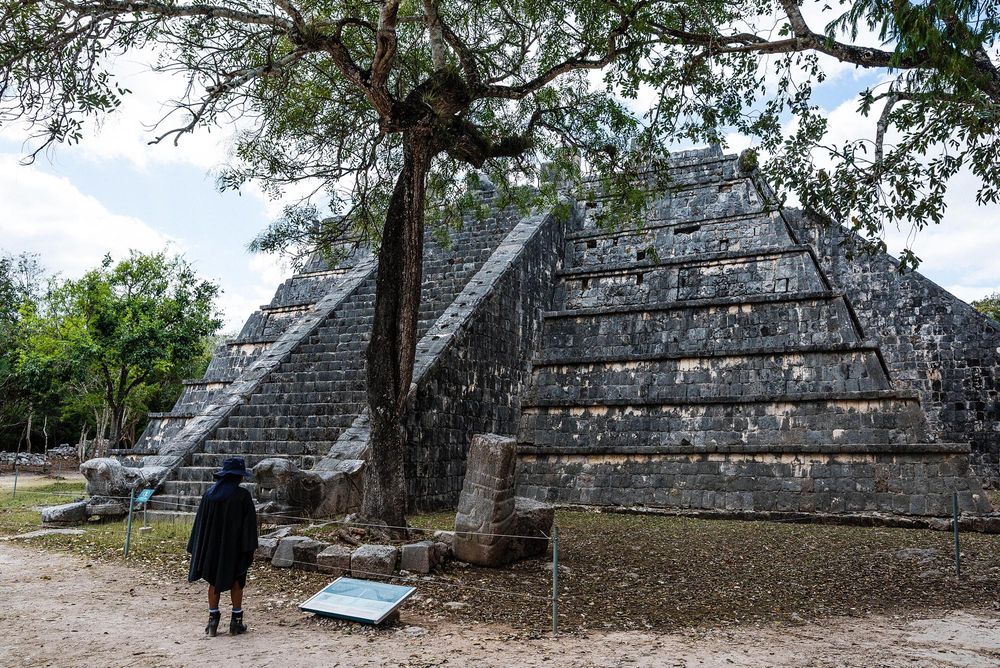 A visitor stands before an ancient Mayan pyramid at Chichen Itza, Mexico, surrounded by trees.