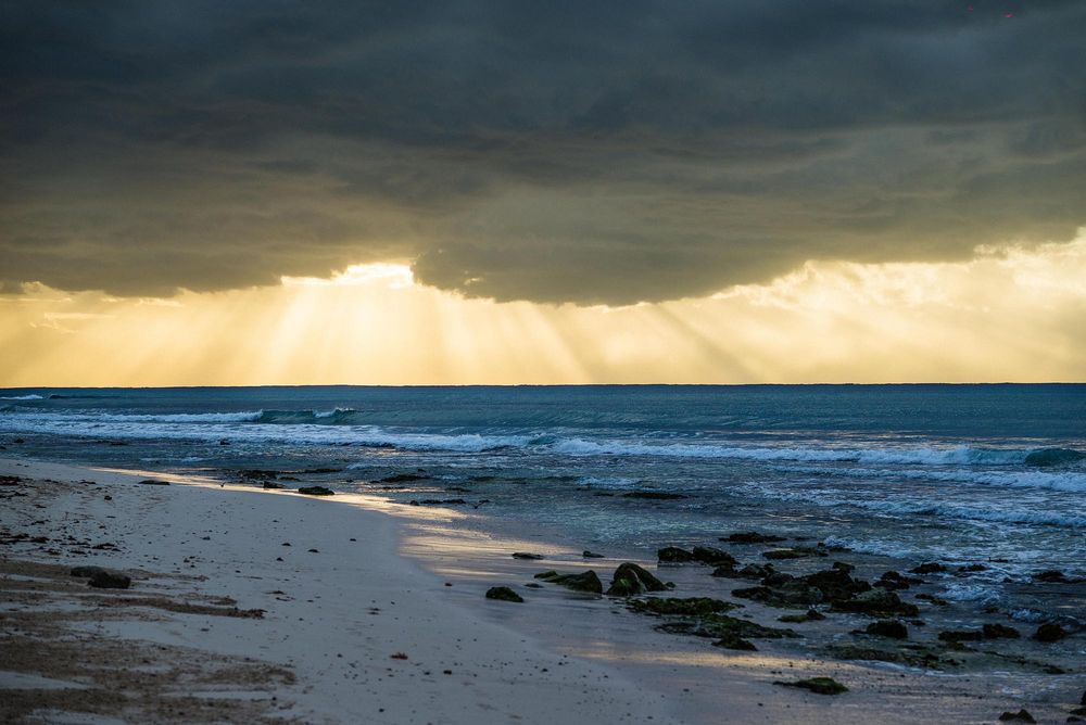 Dramatic beach sunset with golden rays breaking through storm clouds over ocean waves and sandy shore.