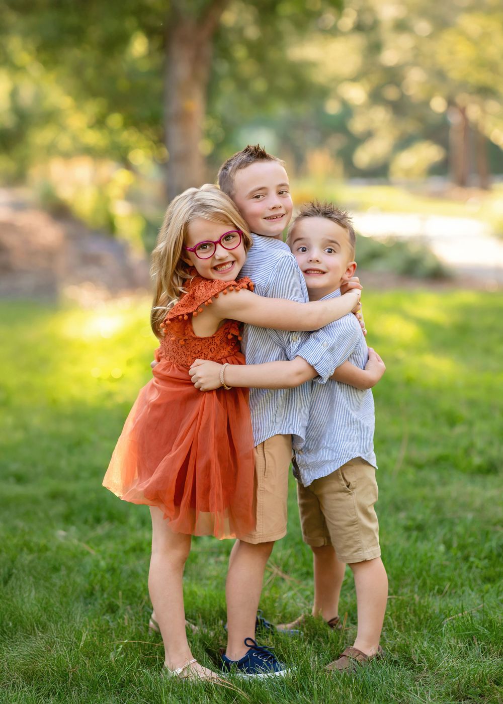 Three young siblings hugging and smiling in a sunny green park.