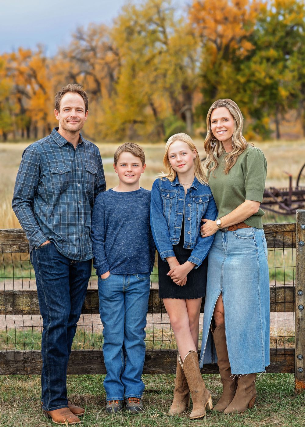 Family of four posing by wooden fence with autumn foliage background.