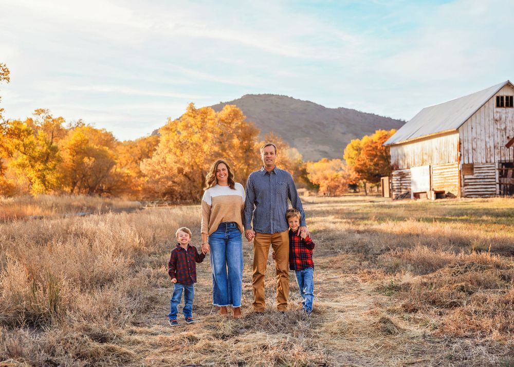Family of four holding hands in golden autumn field near barn.