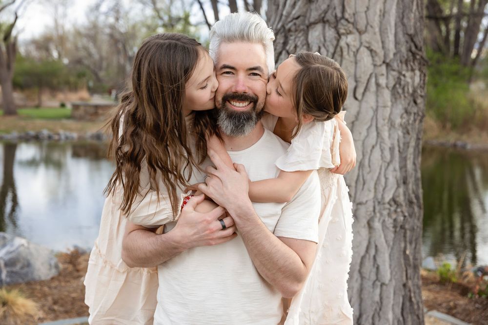 Father being kissed on cheeks by two daughters near a tree outdoors.