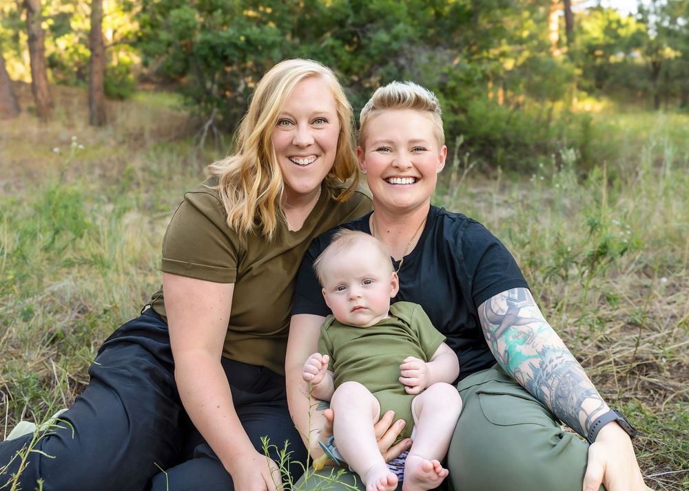Two women smiling with baby in outdoor nature setting.
