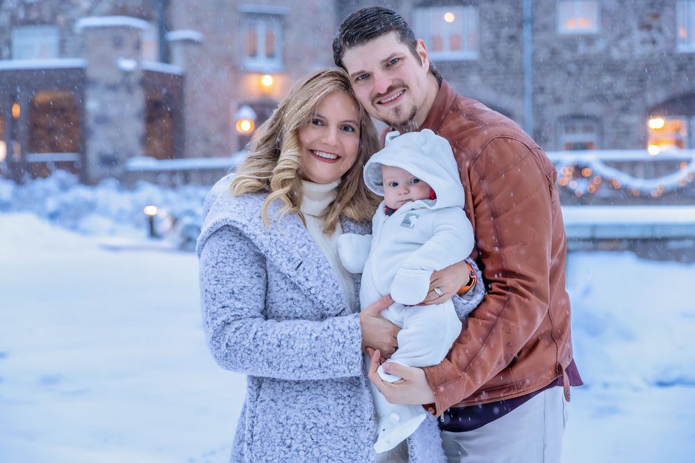 Couple holding newborn baby in snowy winter outdoor setting at dusk.