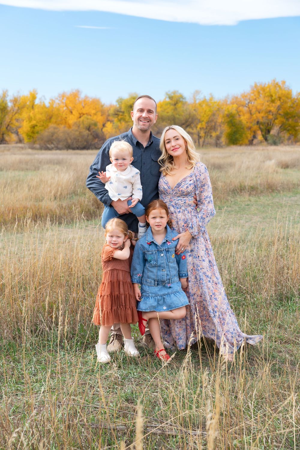 Family of five smiling in golden autumn field under blue sky.